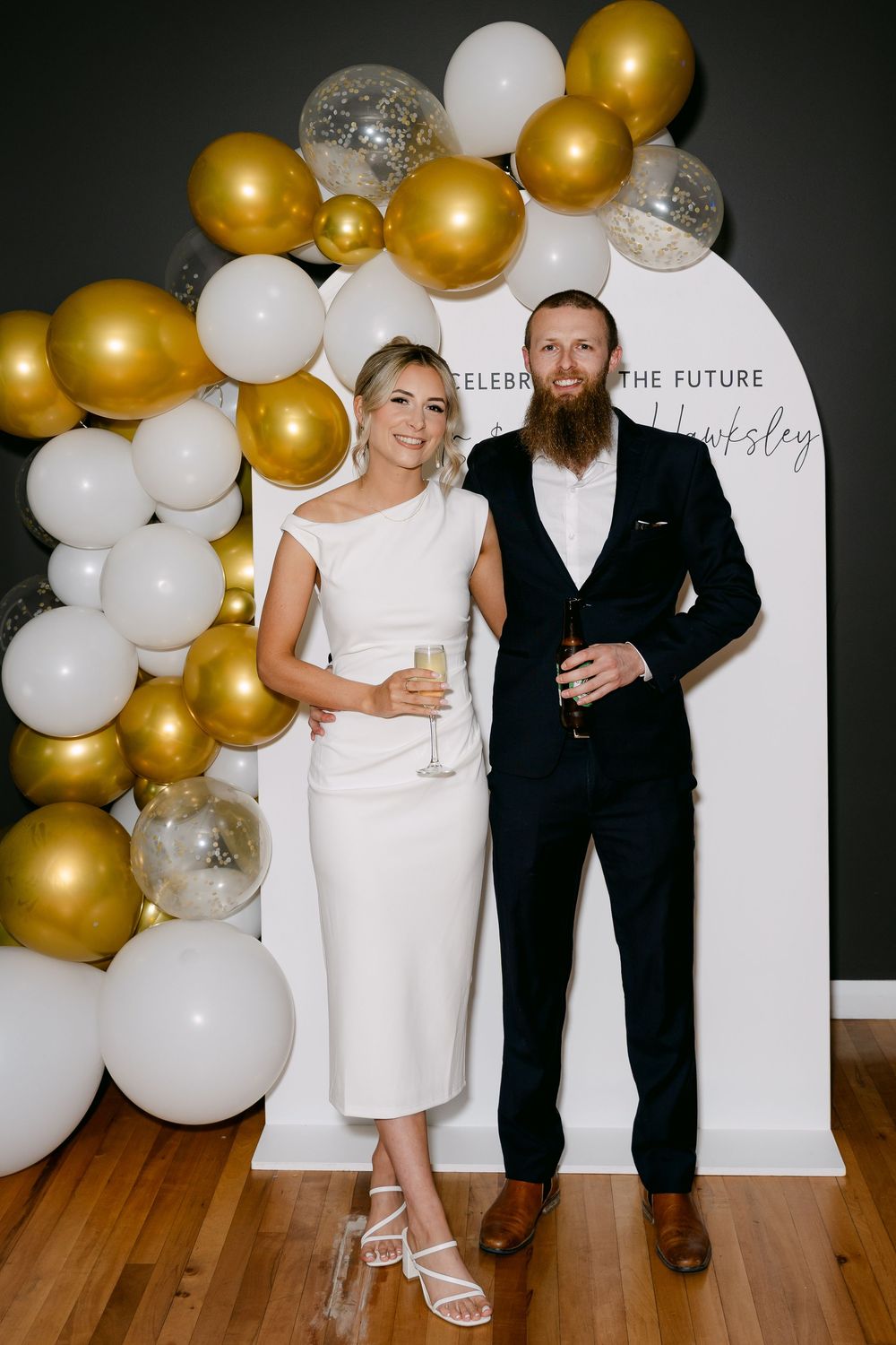 Couple posing at engagement party with gold, white, and silver balloon arch backdrop holding champagne glasses.