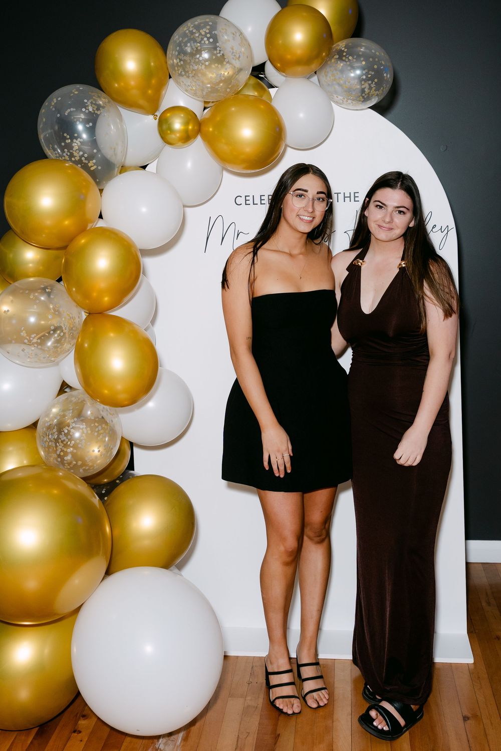 Two women in black dresses posing in front of a white arch backdrop with gold and white balloon garland.