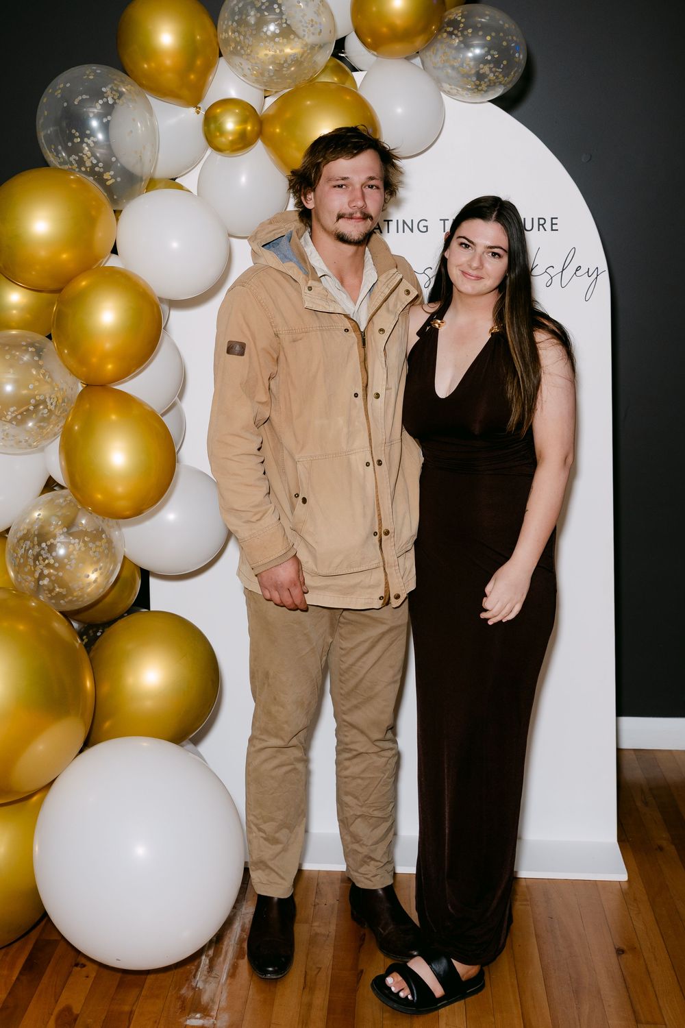 Couple posing together at a celebration with gold, white, and silver balloon garland backdrop.