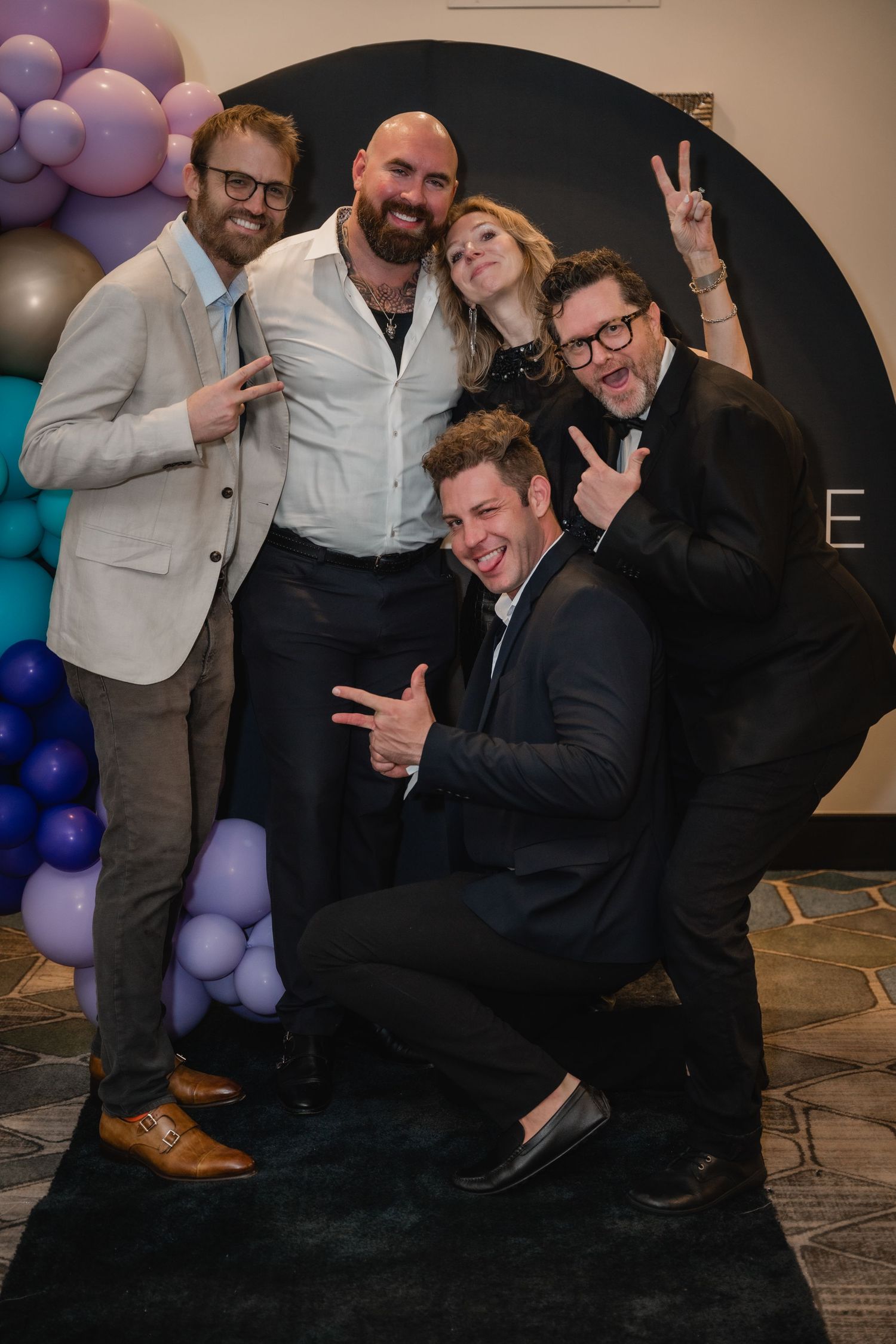 A group poses together for a celebratory photo against a black backdrop with colorful balloon decorations.