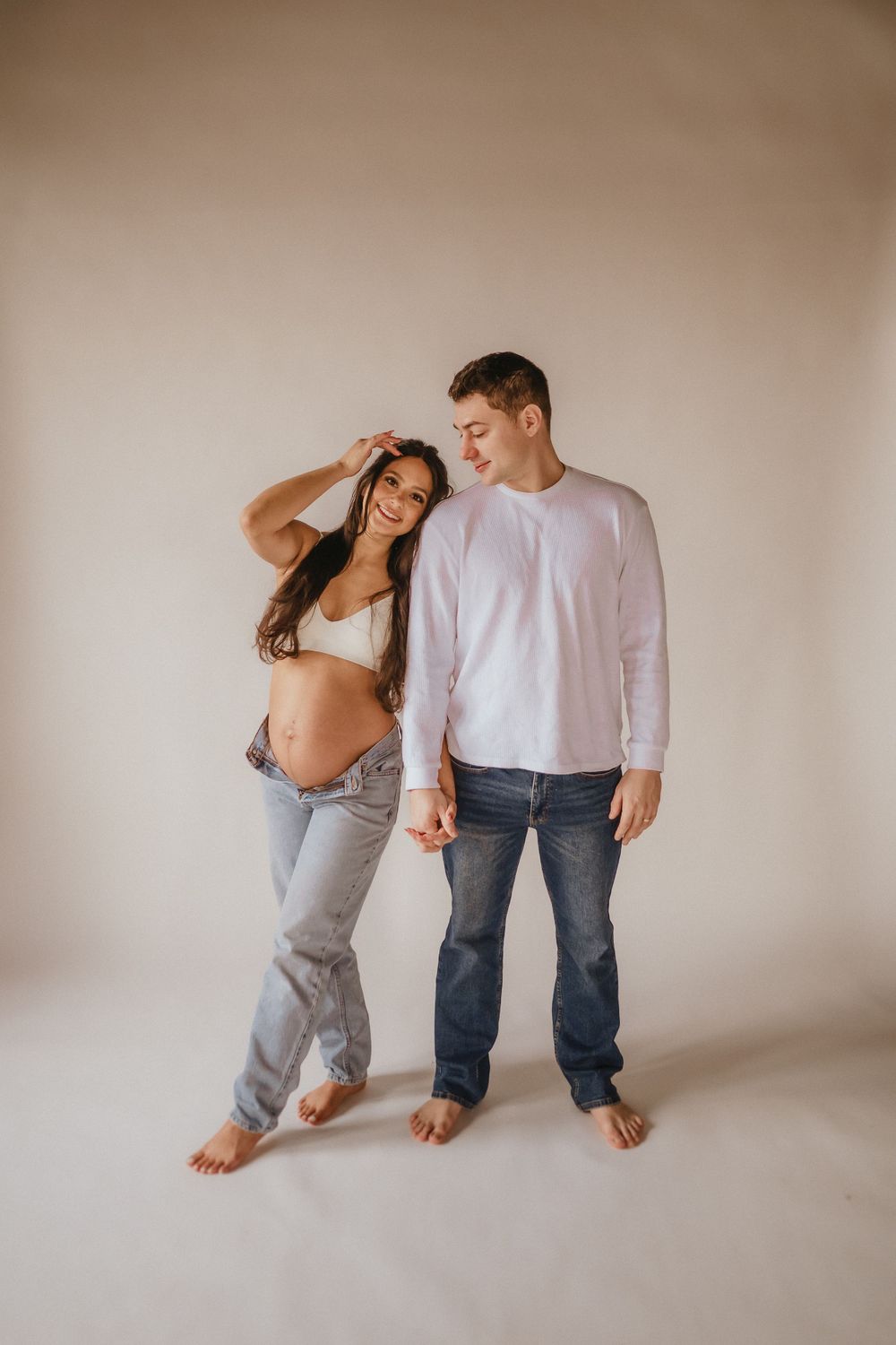 A couple stands barefoot against a neutral background, with the woman wearing a crop top and jeans.