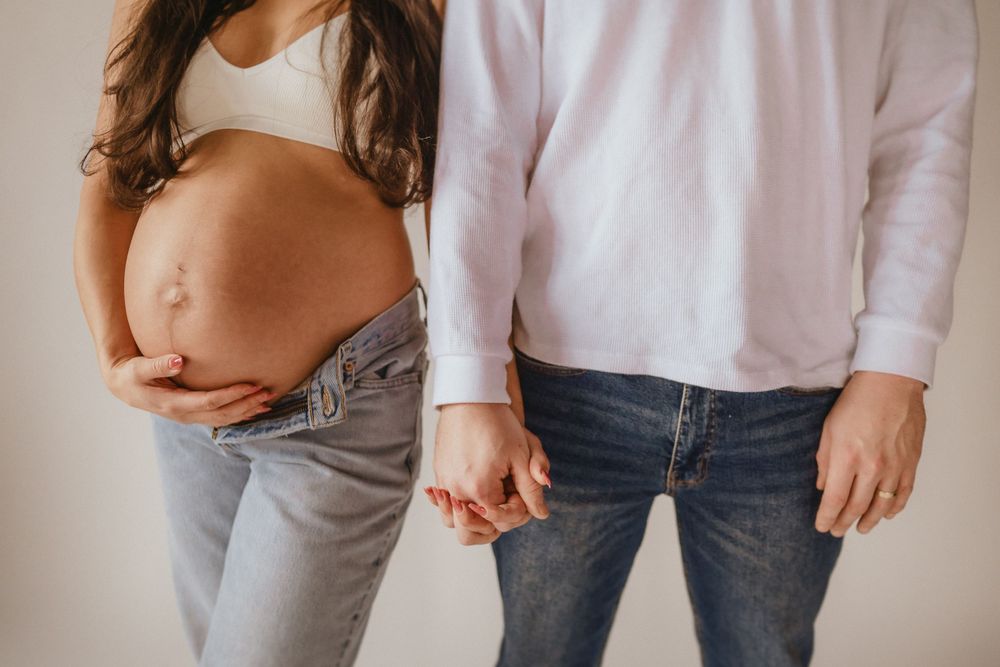 A pregnant woman in jeans and white top holds hands with someone wearing a white shirt against a light background.