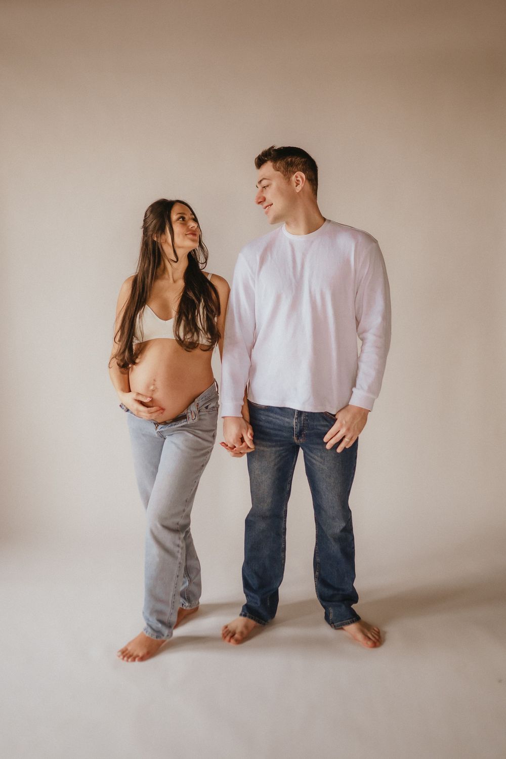 Expectant couple in casual attire posing together against a neutral wall.