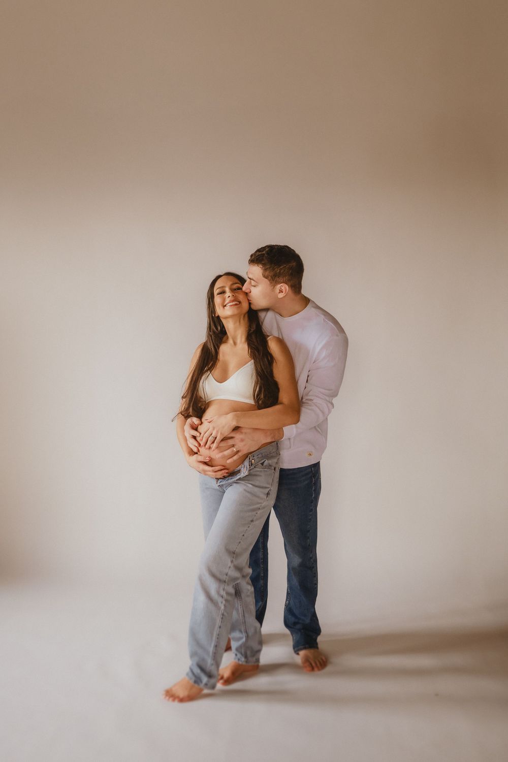 Intimate maternity photo of couple embracing in casual attire against neutral wall in natural light.