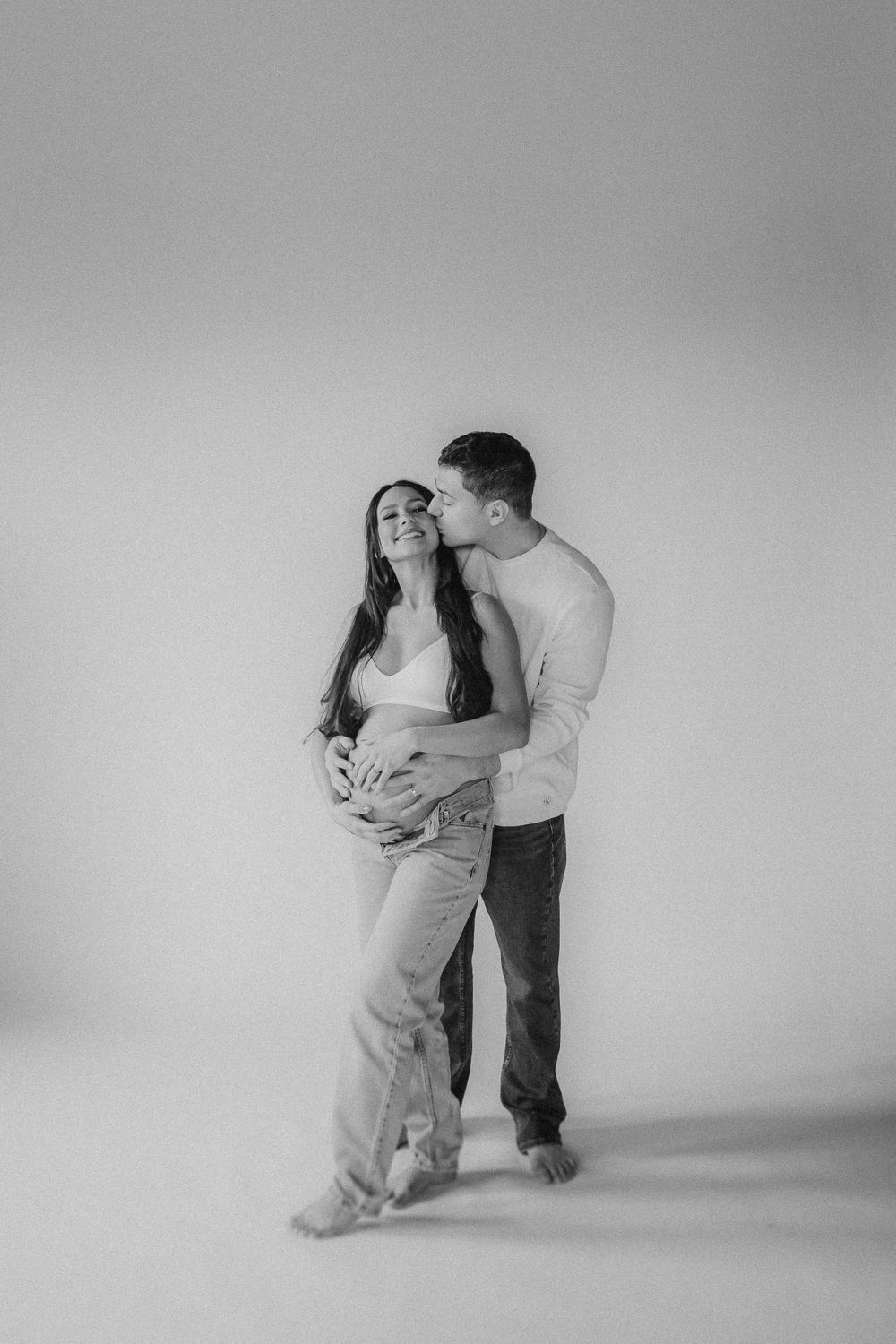 A romantic black and white studio photograph of a couple embracing in casual attire against a light background.