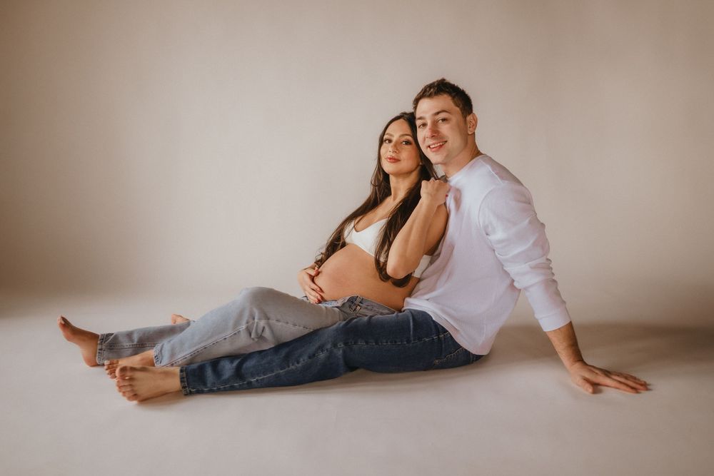 Expecting couple relaxes on floor in casual attire during indoor maternity photoshoot.