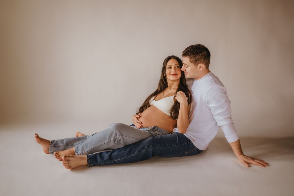 Expecting couple sits intimately on floor during indoor maternity photo session.
