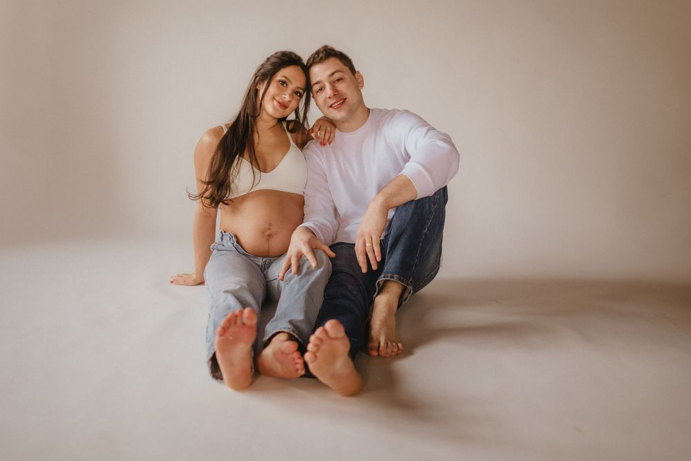 Expectant couple sitting together barefoot on white studio floor during intimate maternity photo session.