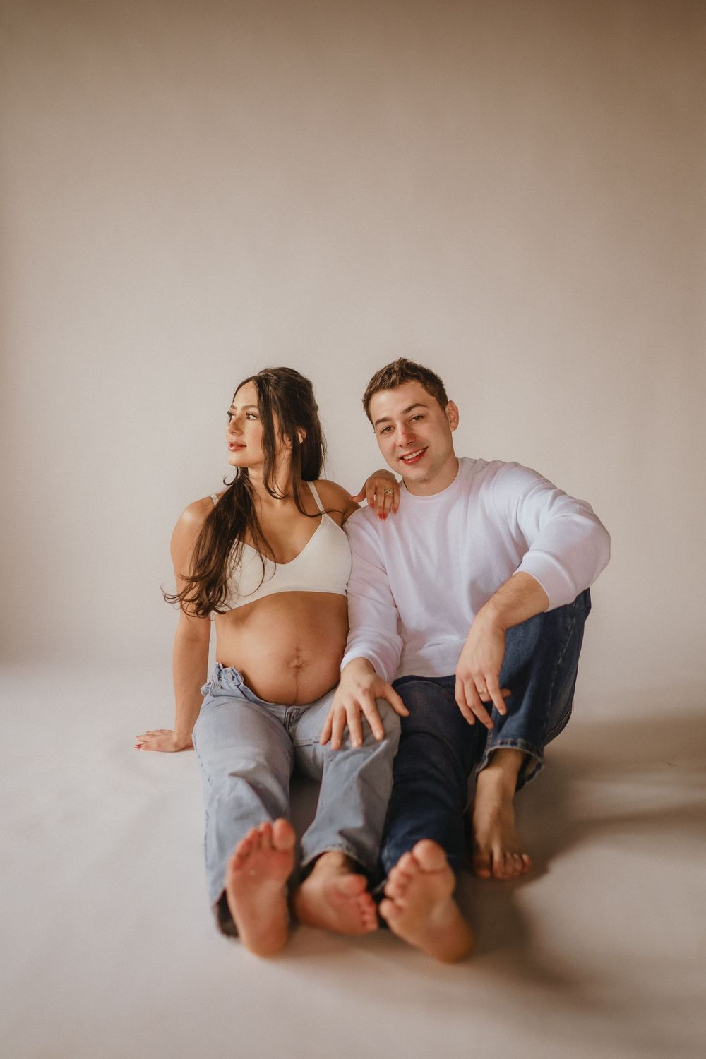 A couple in casual attire sits together barefoot on a light backdrop in a minimalist maternity photoshoot setting.
