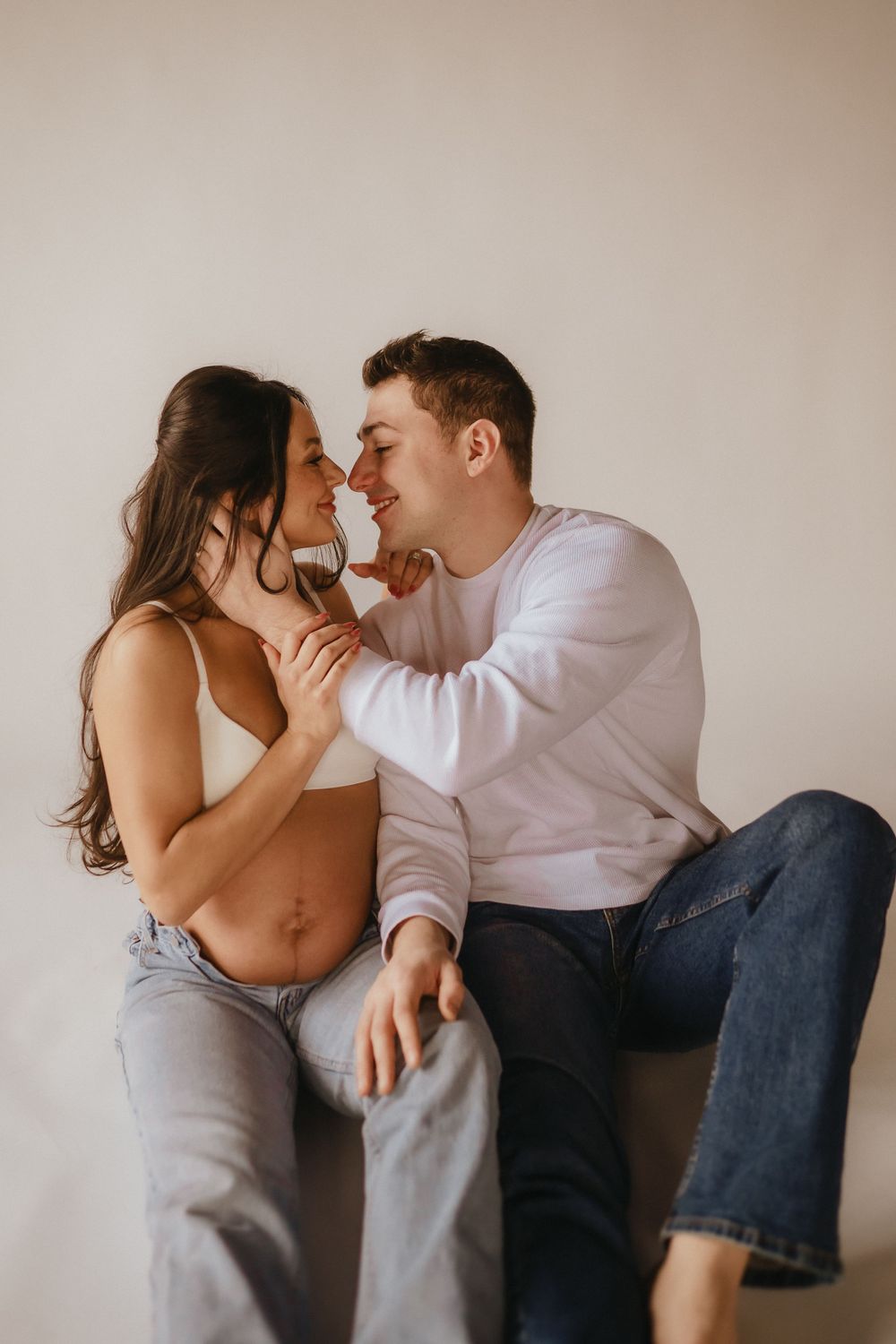 An intimate couple shares a tender moment while sitting together during a maternity photoshoot against a neutral background.