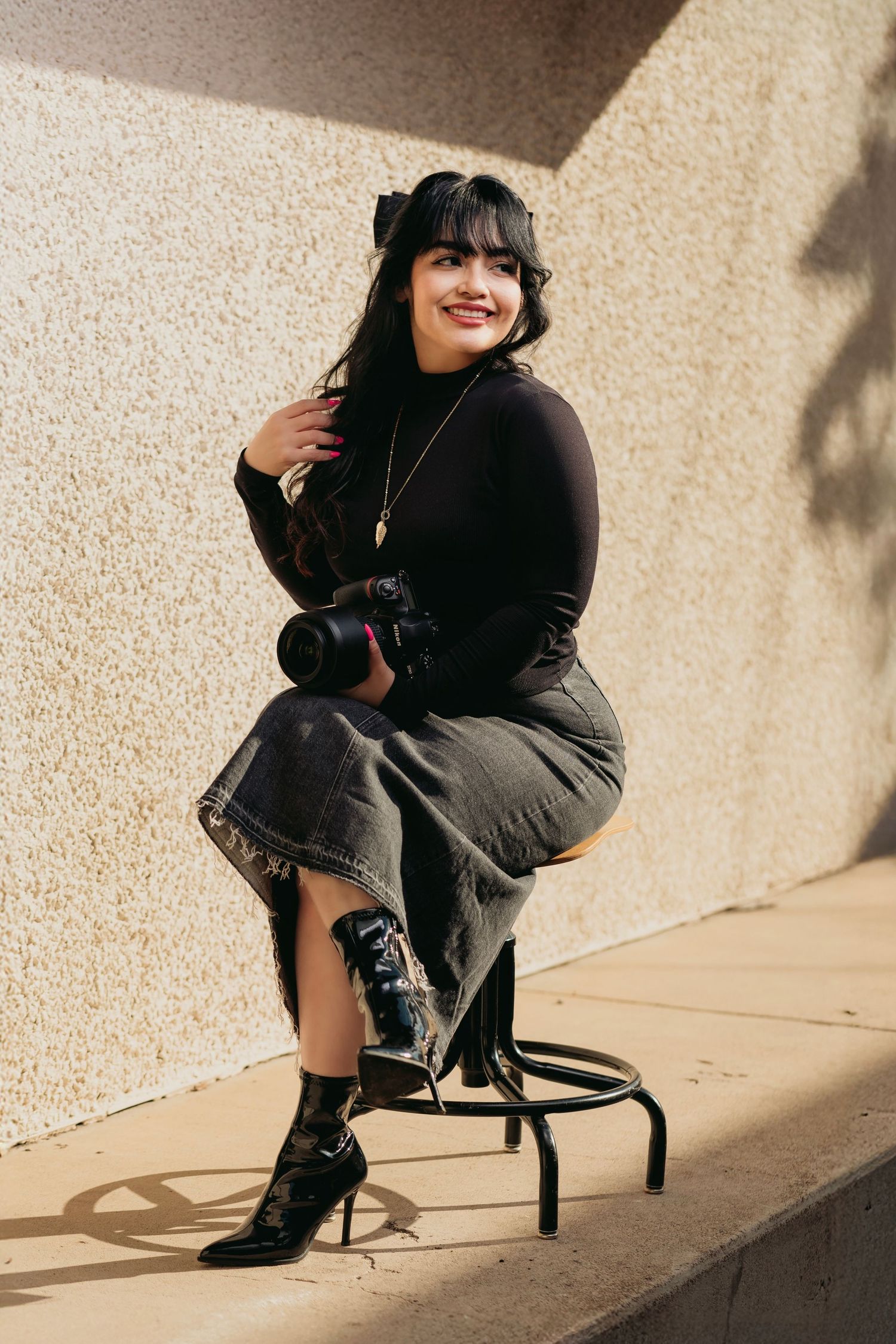 Photographer in black sweater and combat boots sits with camera on stool against beige wall in sunlight.