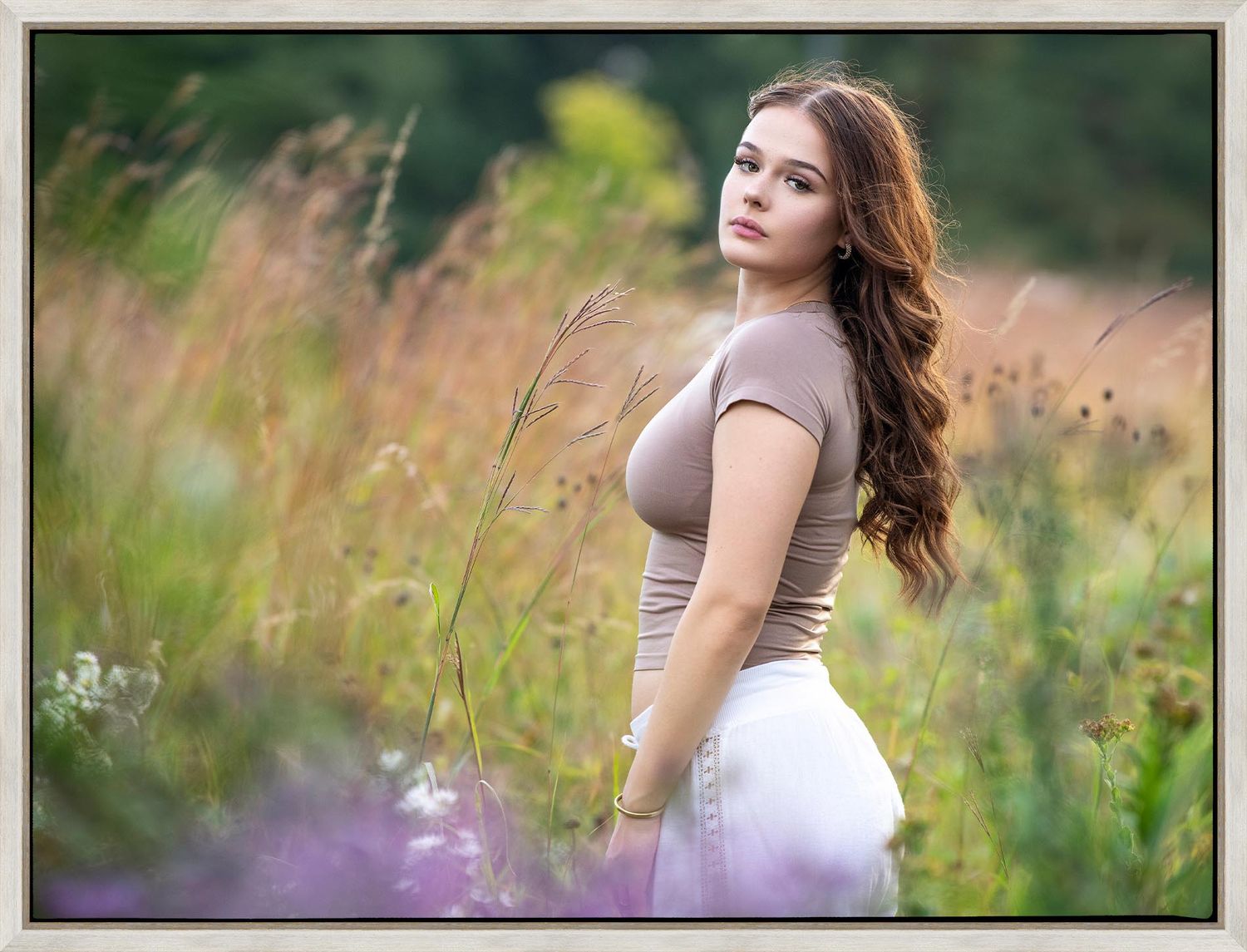 A figure in a beige top and white skirt poses gracefully in a summer meadow filled with wildflowers and tall grass.
