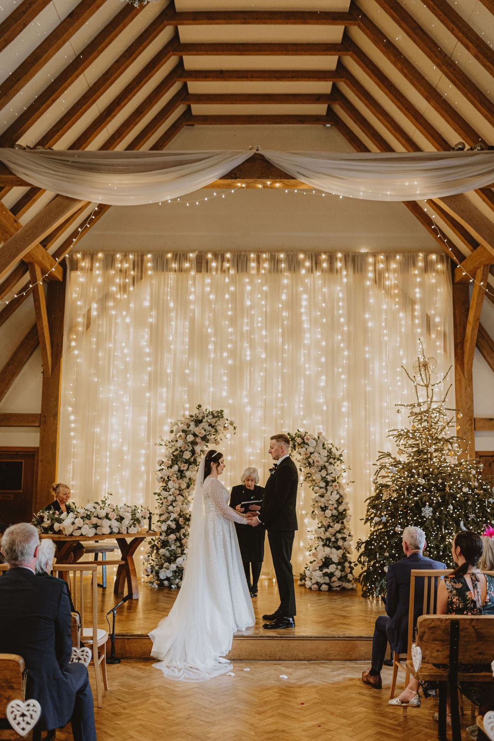 Wedding ceremony setup in a rustic barn venue with white fabric draping and twinkling lights creating a romantic atmosphere.