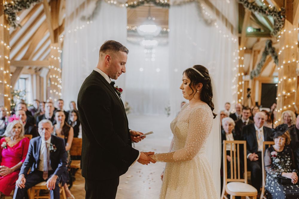Wedding ceremony in progress inside a beautifully decorated barn with fairy lights and floral arrangements.