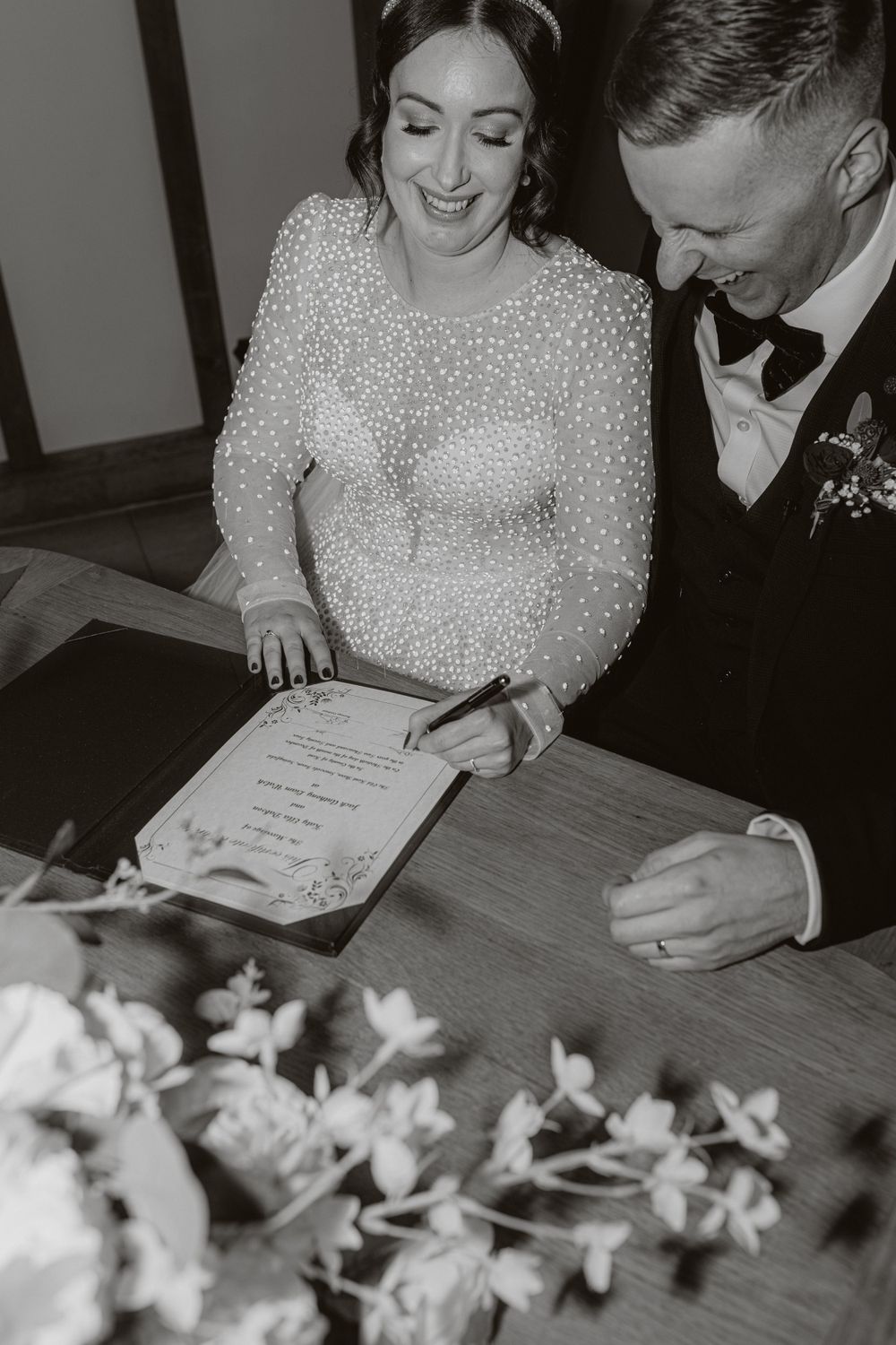 Black and white photo of signing documents at a wedding with floral decorations on wooden table.