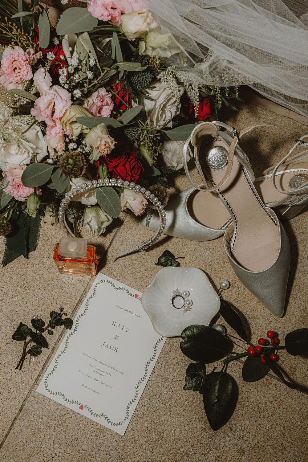 Wedding details including rings, invitations, shoes and pink floral arrangements on beige carpet.