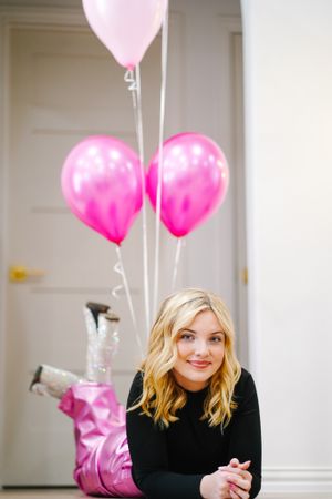 Person lying on white bedding with pink heart-shaped balloons floating above in bright room.