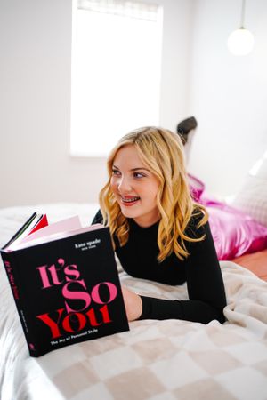 Person in black sweater reading book while lying on white bedding with pink balloons in background.