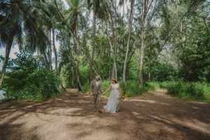 A couple stands together on a dirt path surrounded by tall palm trees and lush tropical foliage in Hawaii.