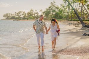 Two people walk together barefoot along a tropical beach with palm trees and ocean waves at sunset.