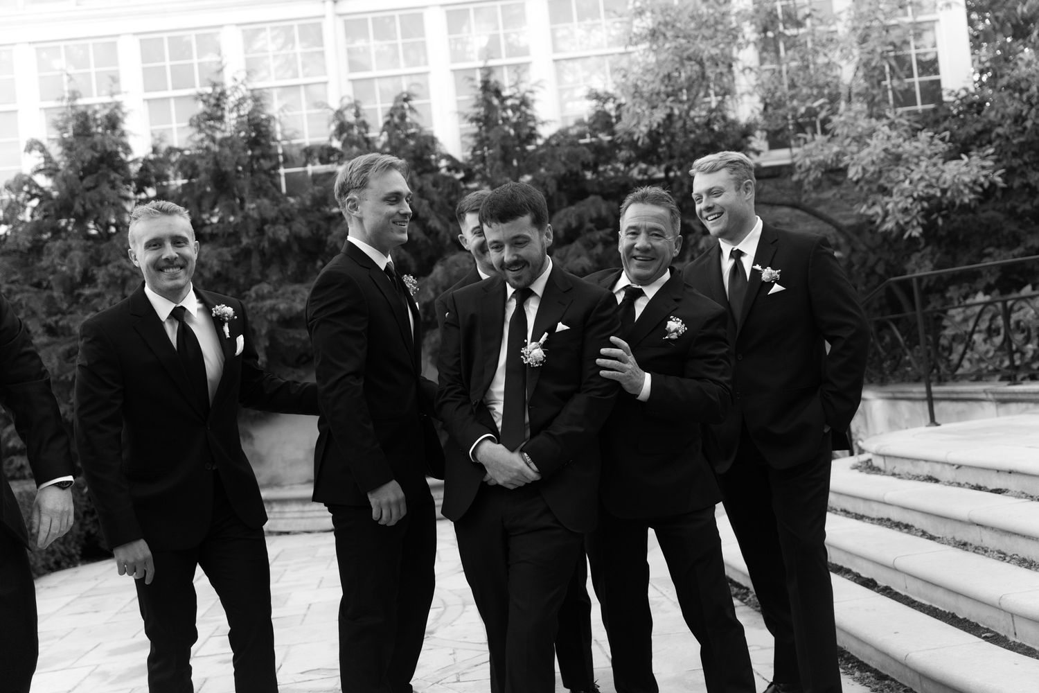 A group of groomsmen in black suits share a candid laughing moment on outdoor stone steps against a leafy backdrop.