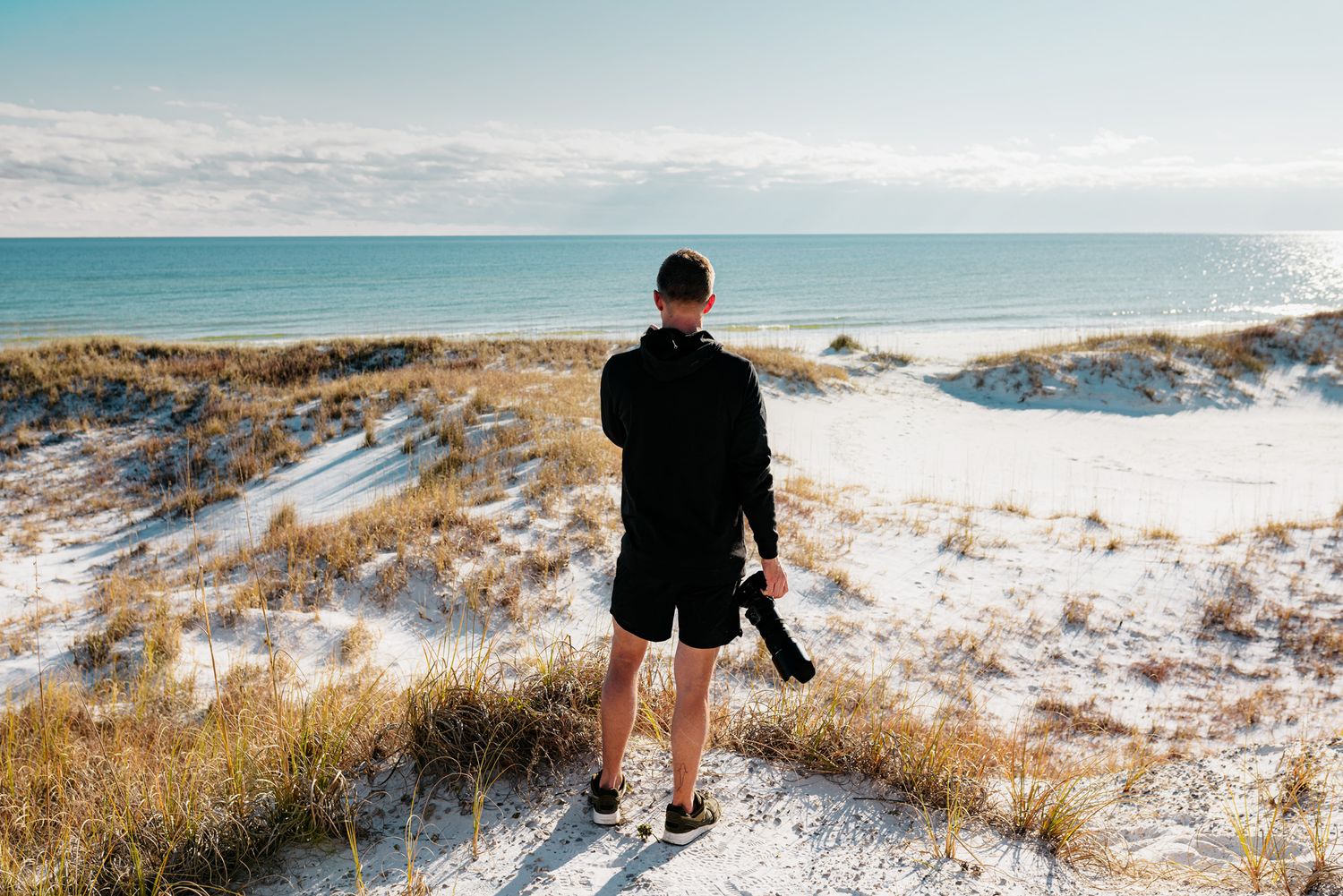 Person in black walking along snowy coastal dunes with ocean view.