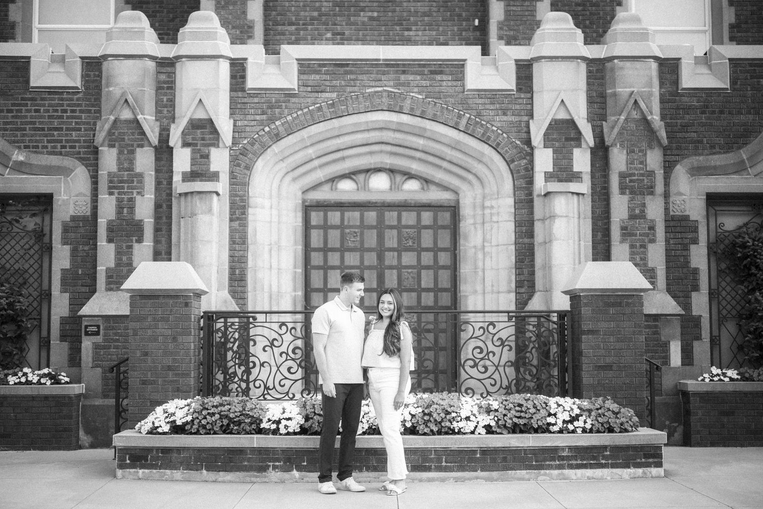A romantic black and white photograph of a couple standing in front of an ornate brick building entrance with gothic details.