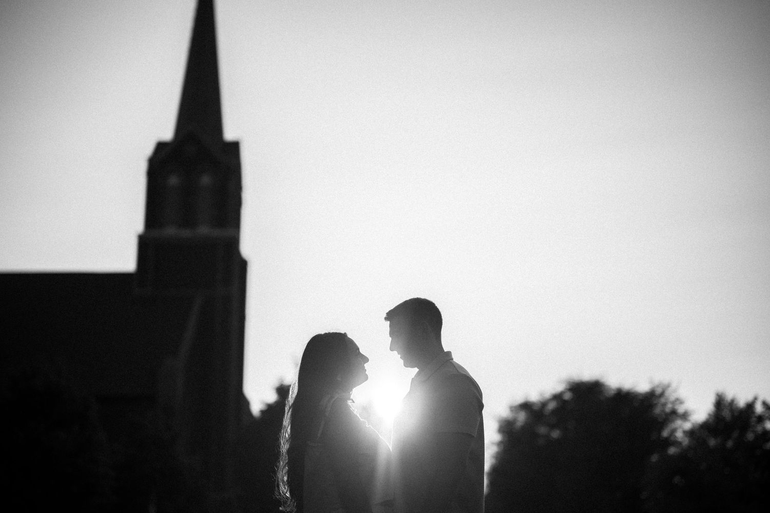 Silhouetted couple in front of church steeple with sunburst between them in romantic black and white composition.
