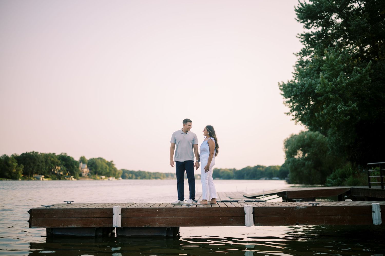 A couple stands together on a wooden dock at sunset overlooking a peaceful lake.