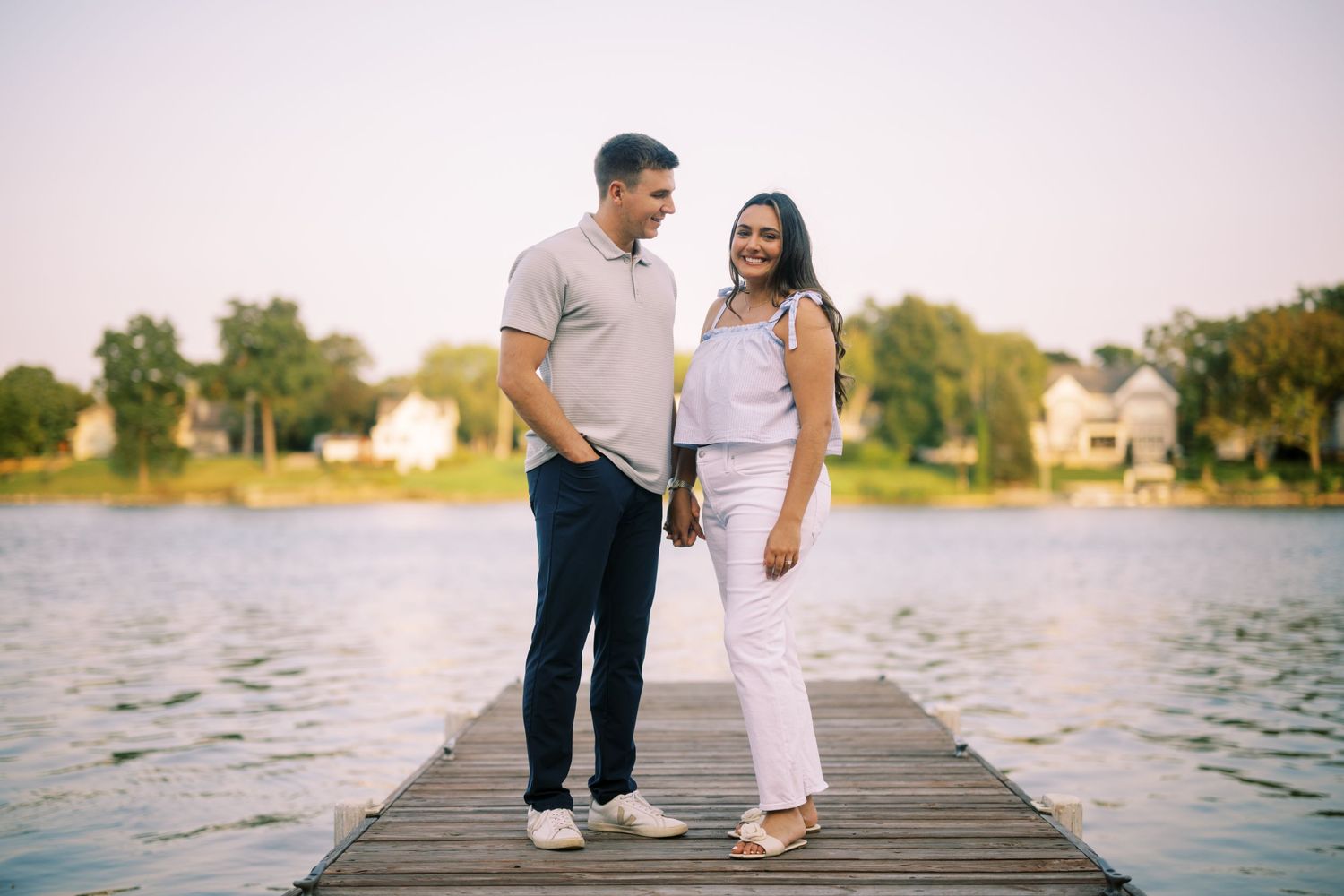 Couple standing together on wooden dock over lake at sunset wearing matching white and light colored outfits.