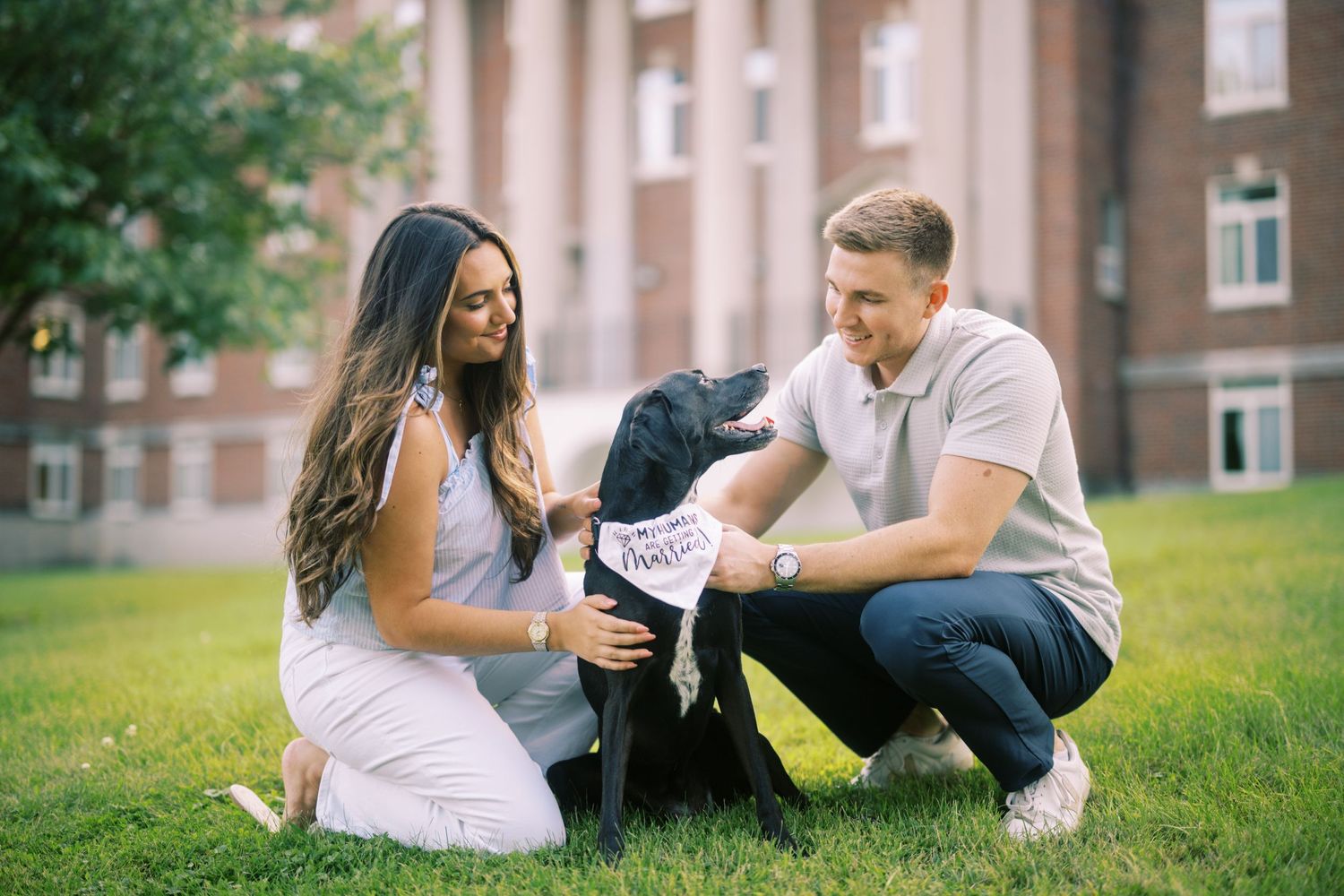 A couple pets a black dog on green grass outside a brick building on a sunny day.