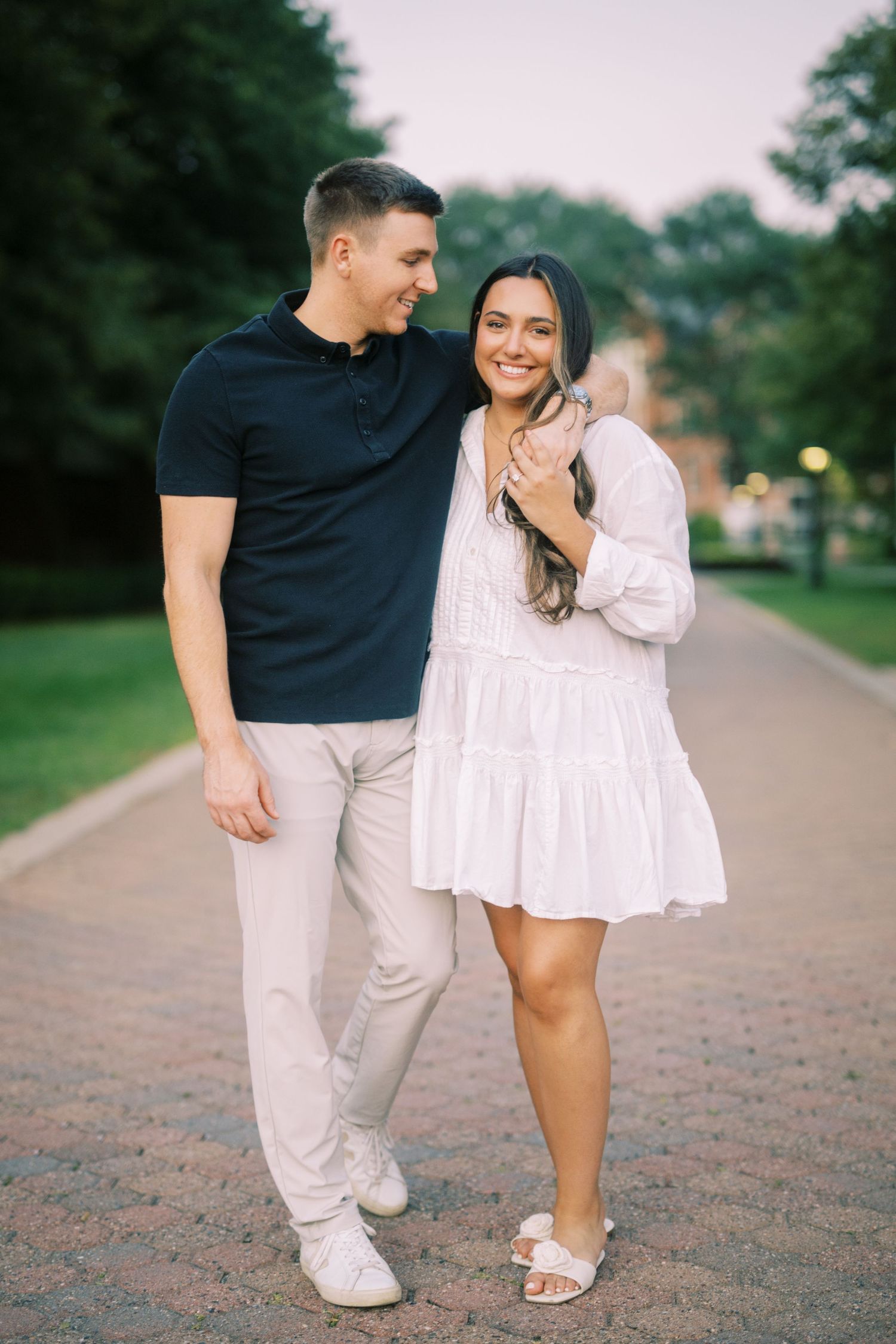 A couple walking together on a brick path in a park, wearing casual summer outfits against a green background.