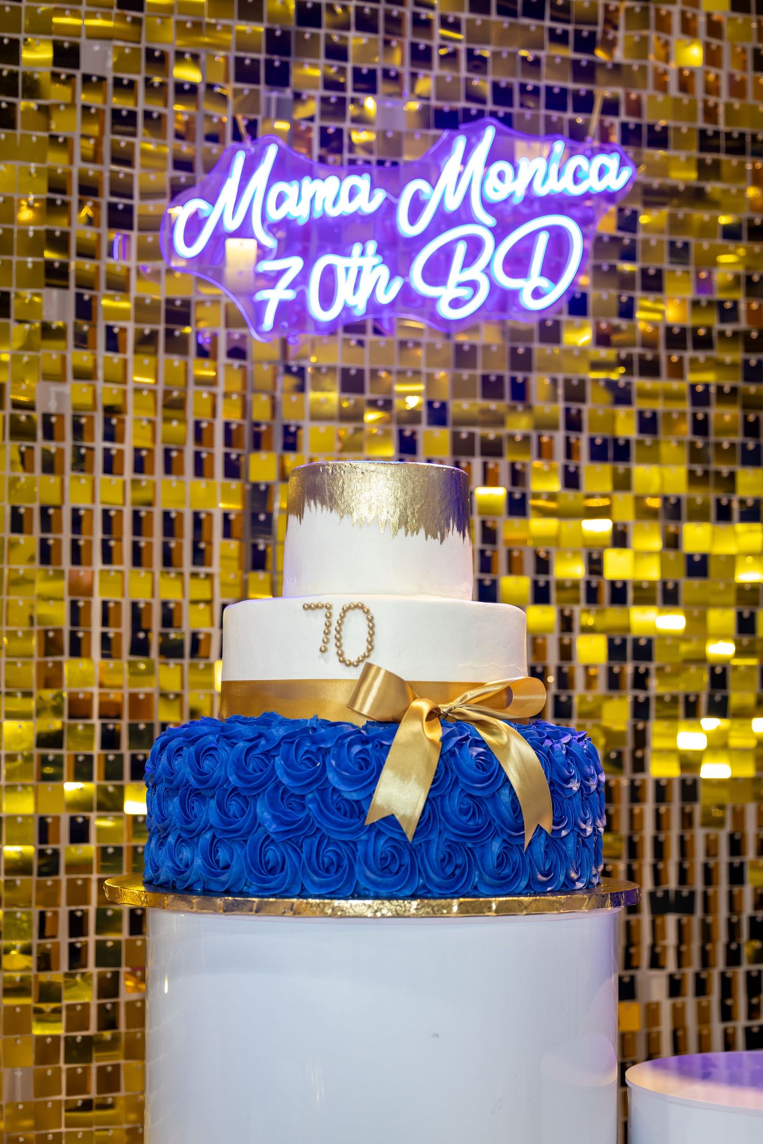 Three-tiered birthday cake with royal blue rosettes and gold accents against a golden mosaic wall.