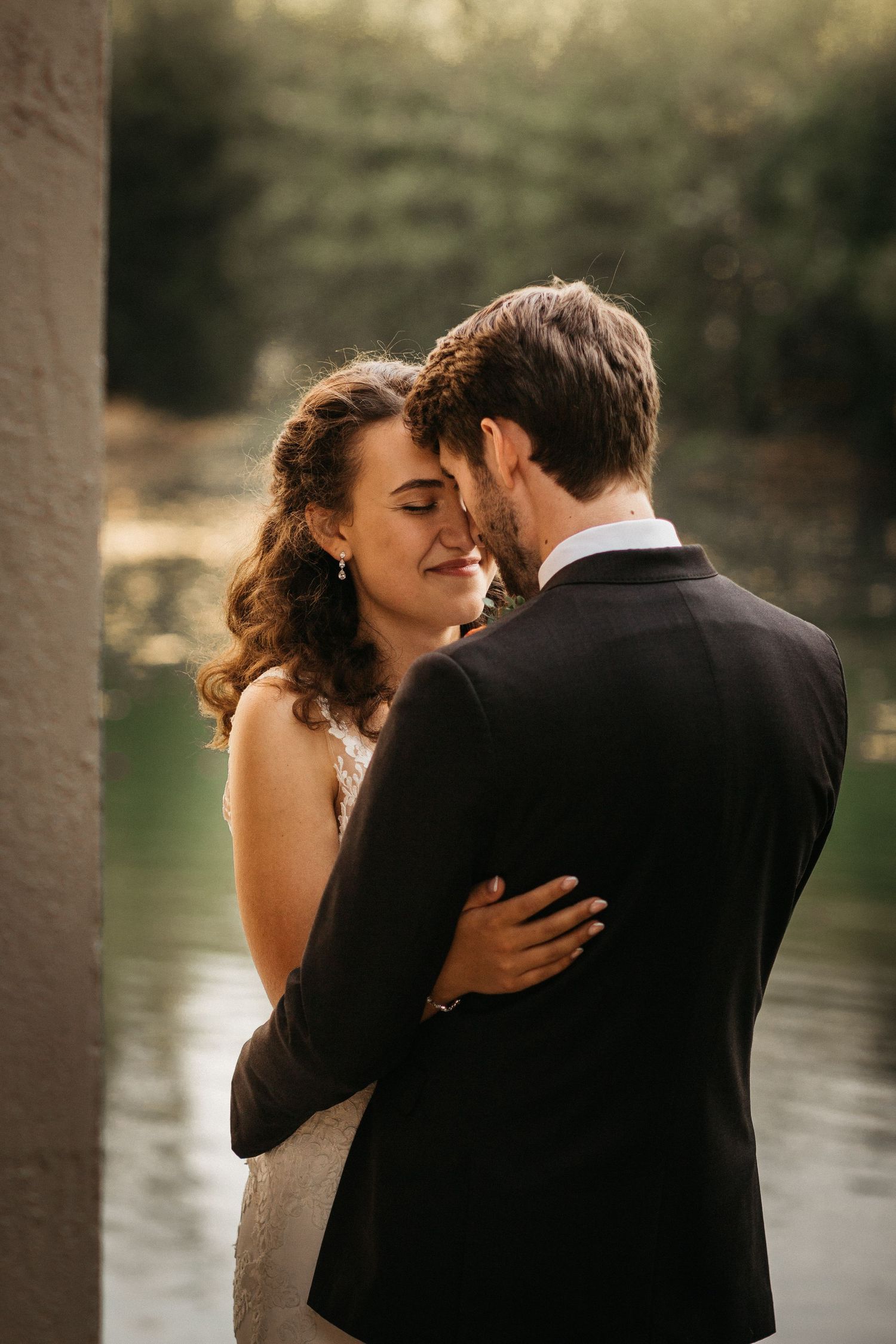 A romantic wedding moment captured at sunset by a lake with a couple embracing in formal attire.