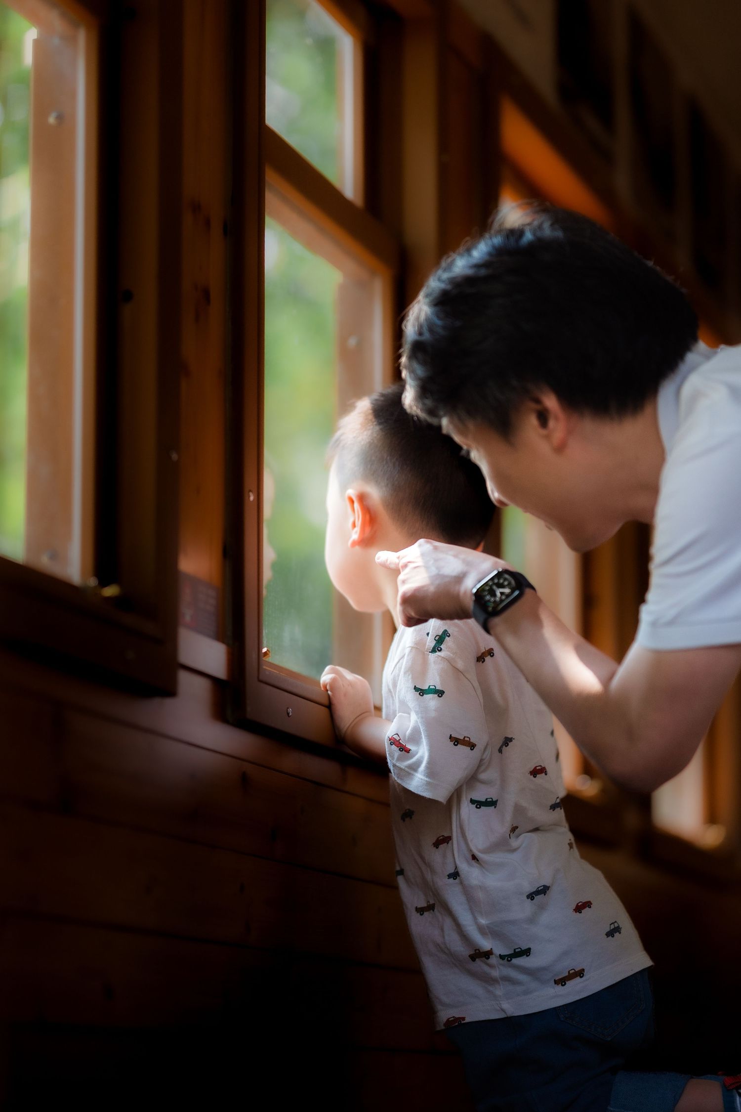 A father and young child look out a wooden cabin window together, sharing a curious moment in warm natural light.