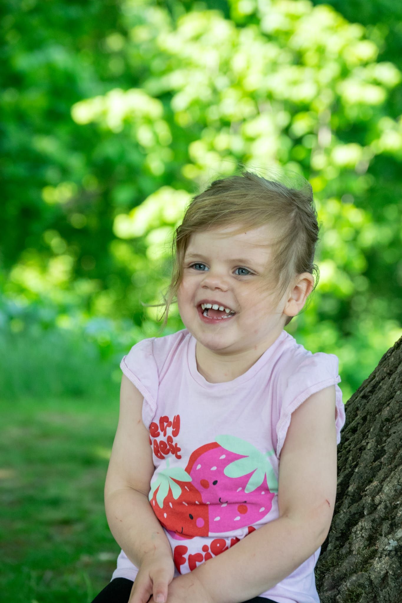 Happy smiling toddler girl in pink strawberry shirt leaning against a tree outdoors.