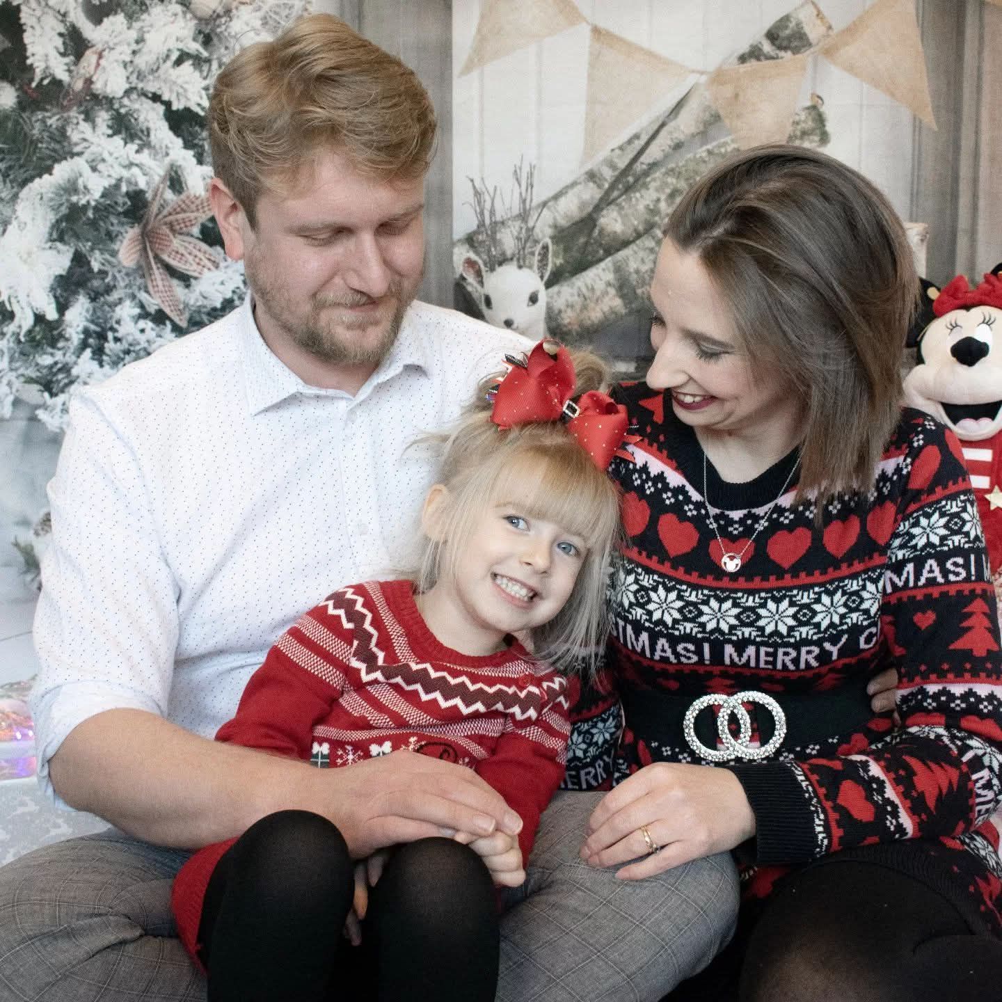 Happy family of three wearing matching Christmas sweaters posing in front of a festive tree.