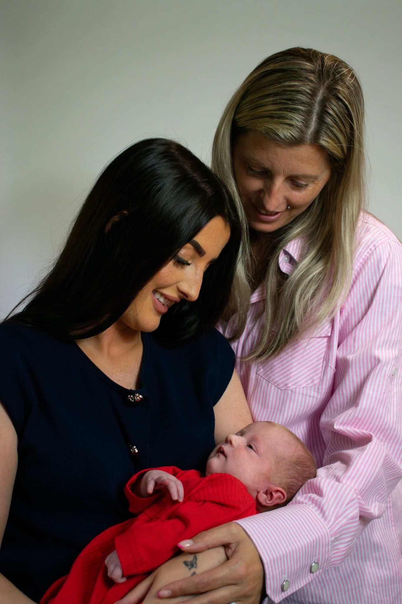 Two smiling women lovingly admiring a newborn baby wrapped in a red blanket.