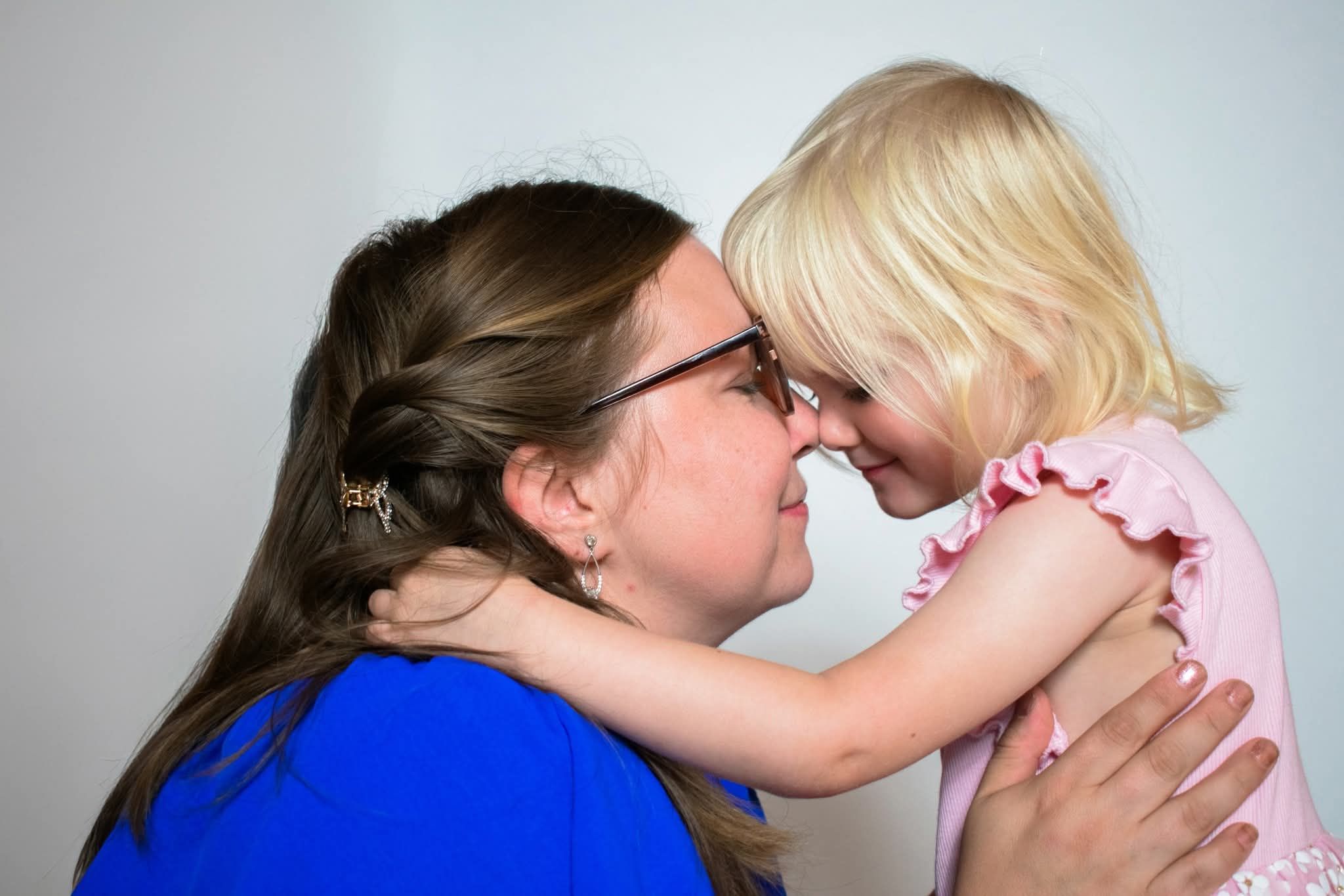 A mother touching foreheads and smiling with her young blonde daughter in a pink dress.