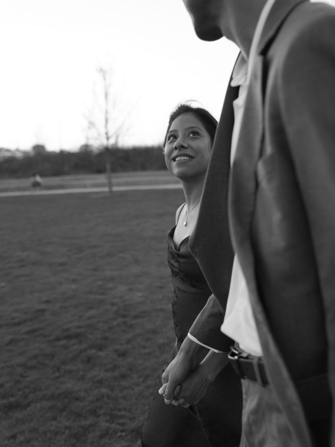 Black and white photo of a smiling woman holding hands with a man in a suit outdoors in a park.