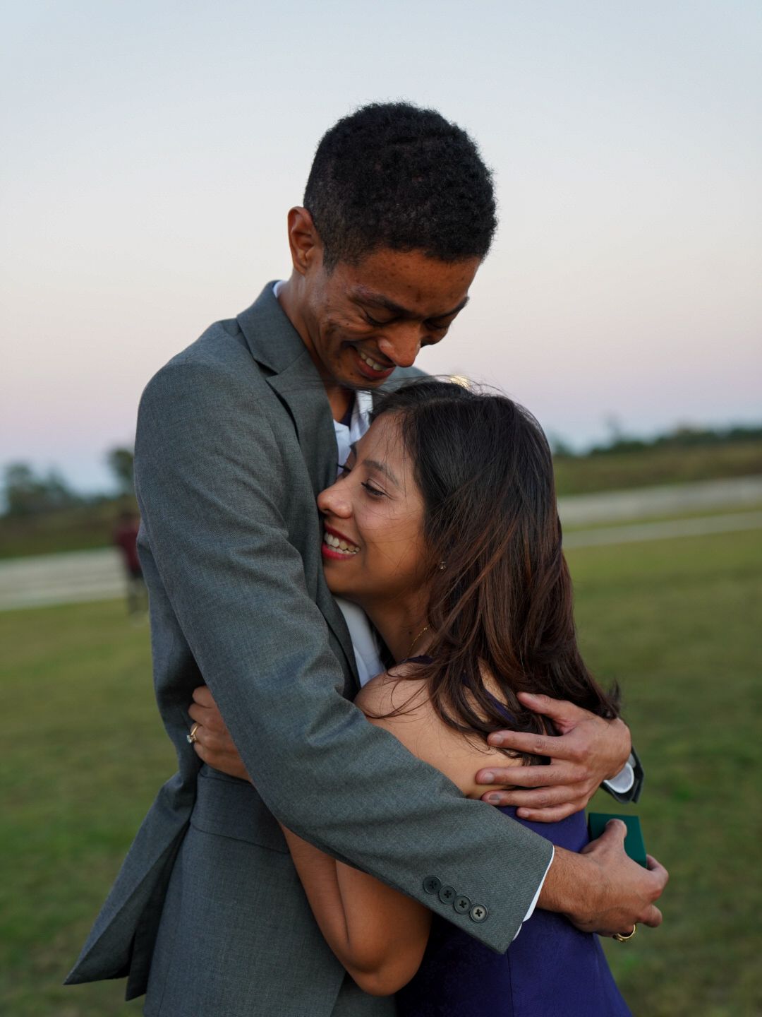 Happy couple embracing and smiling outdoors at dusk, man in gray suit hugging woman in purple dress on grassy field.