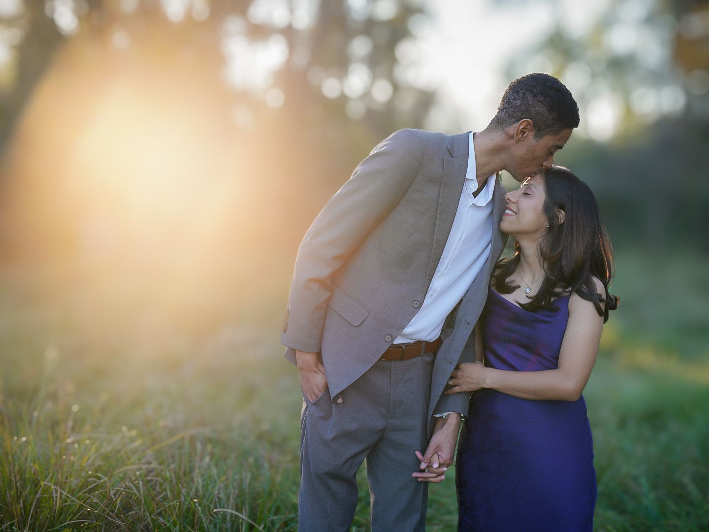 Couple sharing a romantic moment at sunset in a green field, man in tan blazer and woman in purple dress.