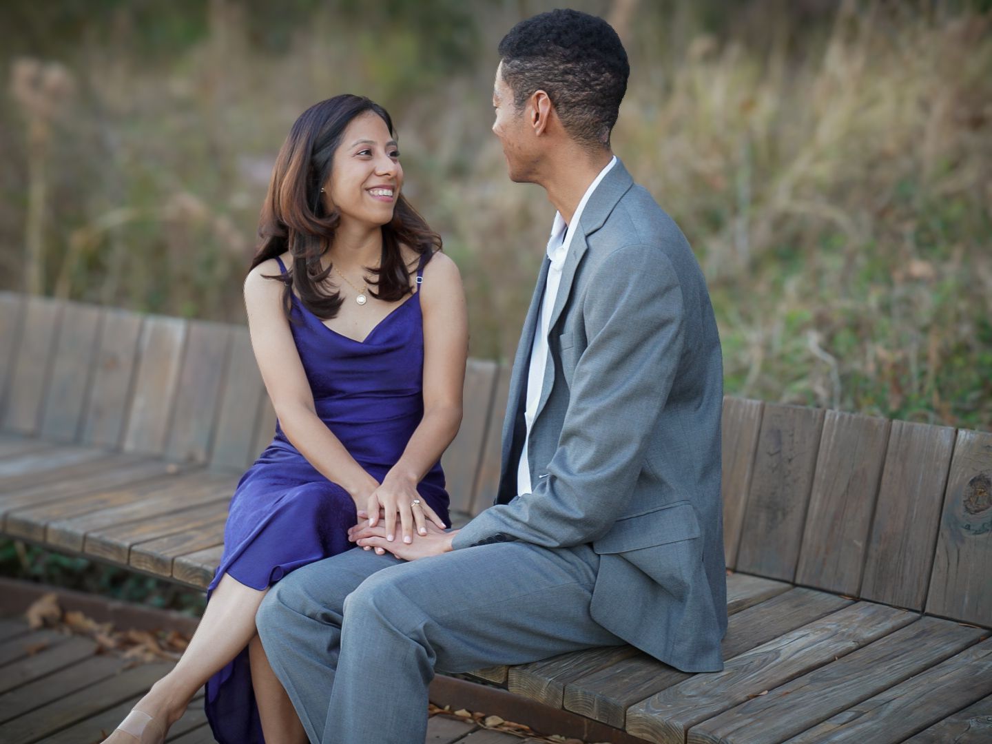 Couple sitting on a wooden bench outdoors, woman in purple dress and man in gray suit holding hands and smiling.