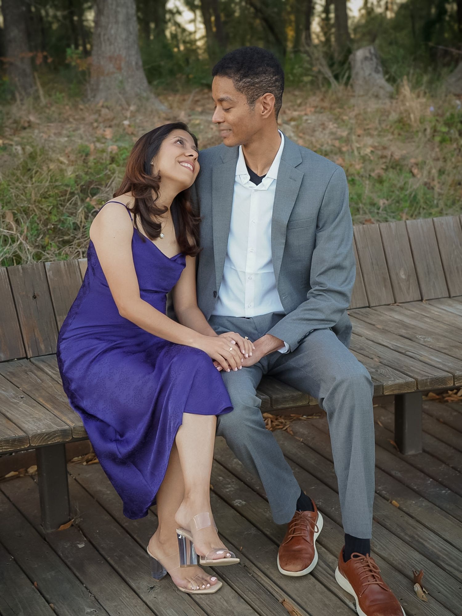 Couple sitting on wooden bench holding hands, woman in purple dress and man in gray blazer smiling at each other outdoors.