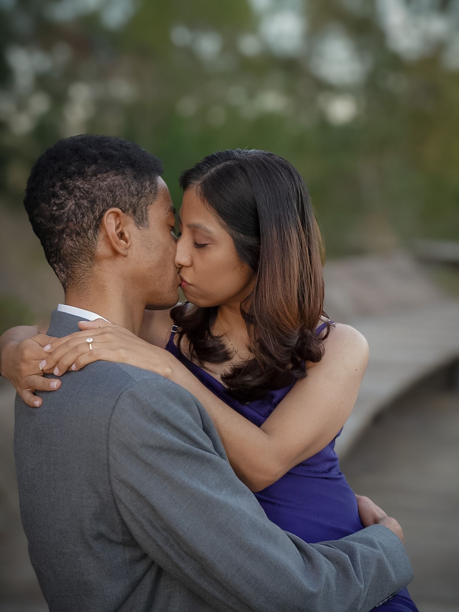 A couple shares a romantic kiss outdoors, the woman in a purple dress embracing a man in a gray suit.