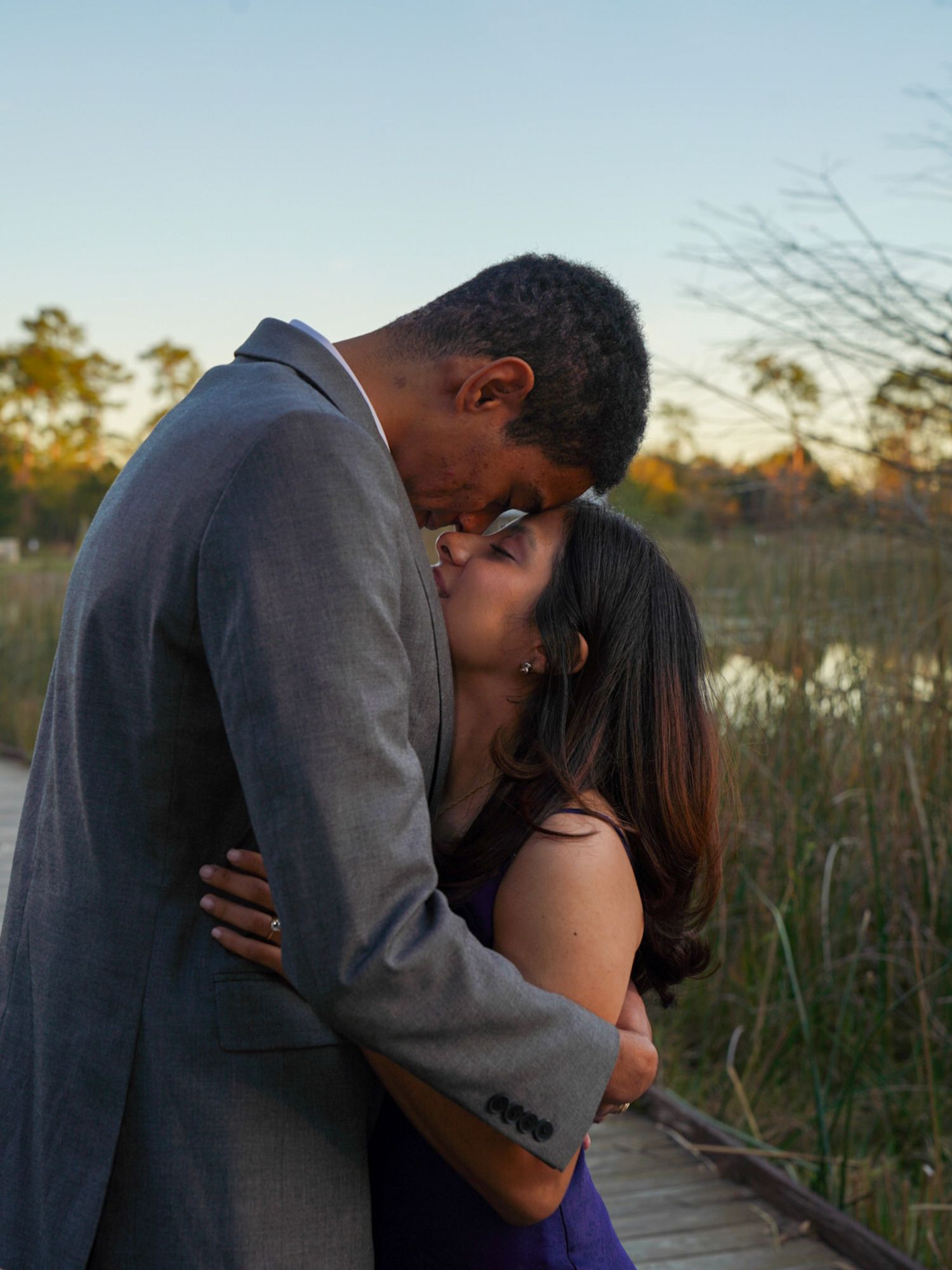 Couple sharing a romantic kiss outdoors near a marsh at sunset, man in gray suit embracing woman in purple dress.