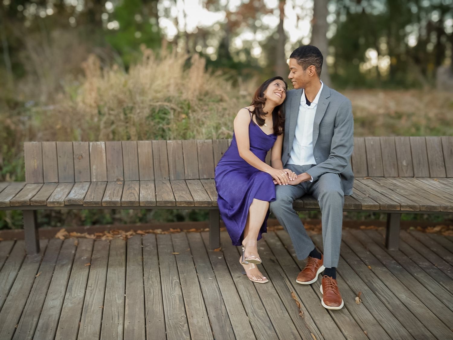 Couple sitting together on a wooden bench in a park, woman in purple dress and man in gray suit holding hands.