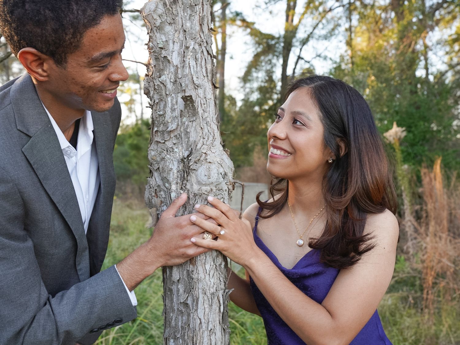 Smiling couple holding hands around a tree trunk outdoors, woman in purple top and man in gray blazer.