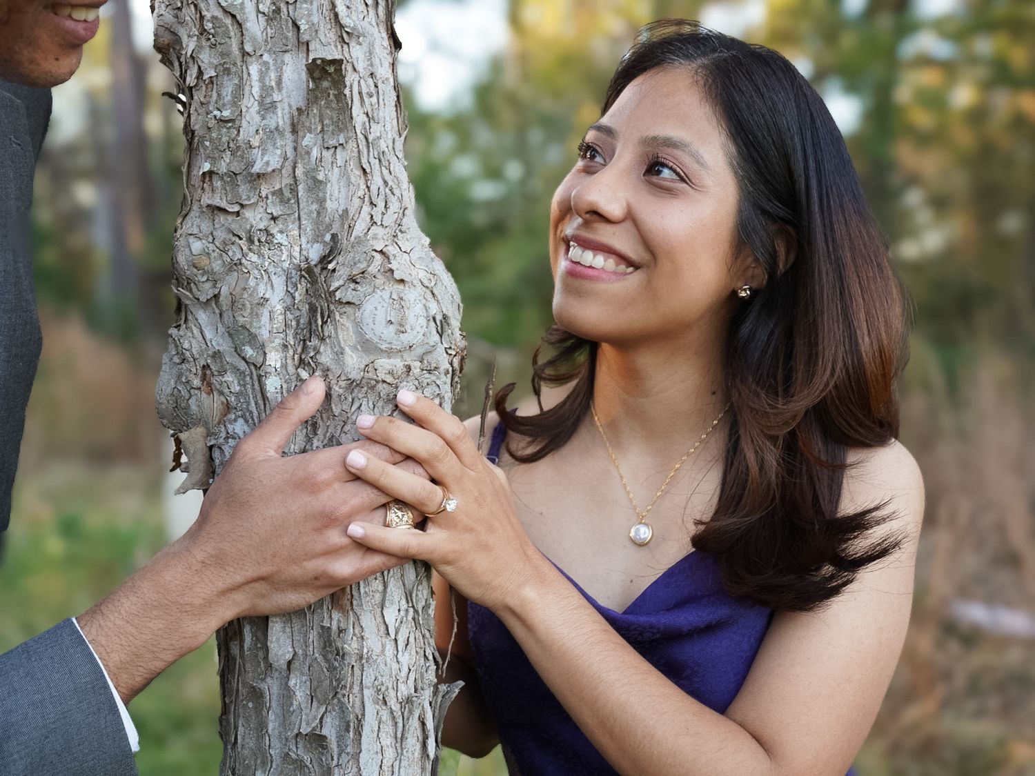 Smiling woman holding hands with partner near a tree trunk in an outdoor setting.