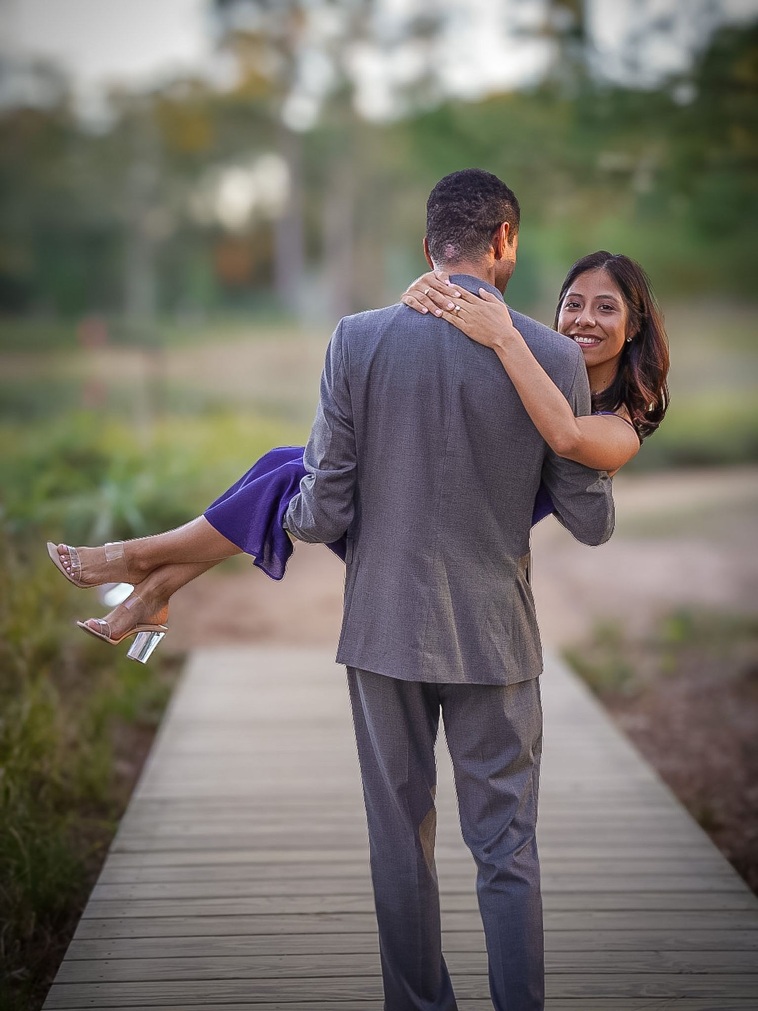 A man in a gray suit carries a smiling woman in a purple dress along a wooden boardwalk.
